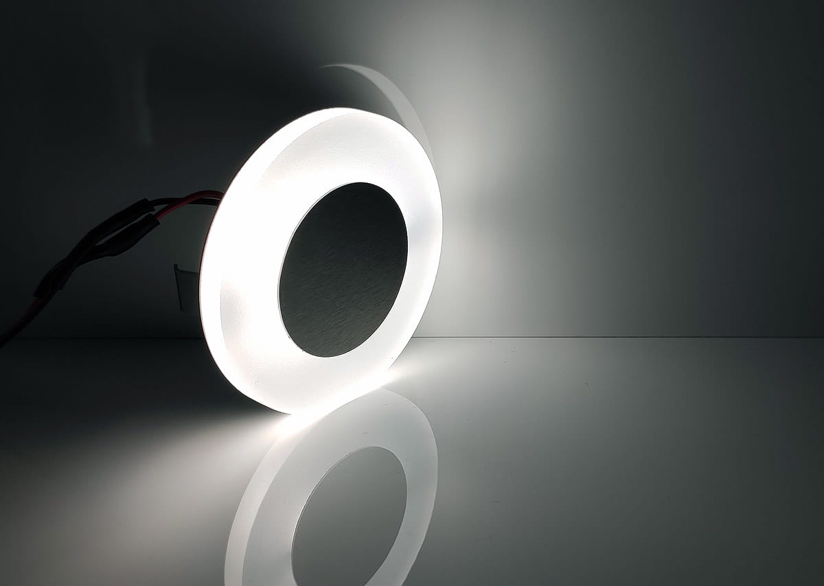 Oti LED oprawa schodowa 230V w kolorze białym, barwa zimna 6500K. Okrągły kształt z matowym wykończeniem zewnętrznym i czarnym wnętrzem. W ujęciu z boku, z przewodami elektrycznymi w tle, na błyszczącej powierzchni.