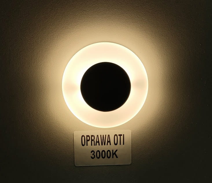 Oti LED oprawa schodowa w kolorze białym, z okrągłą ramką emitującą ciepłe światło (3000K). Widoczna jest czarna środkowa część oraz tabliczka z napisem OPRAWA OTI 3000K. Produkt zamontowany na ścianie.