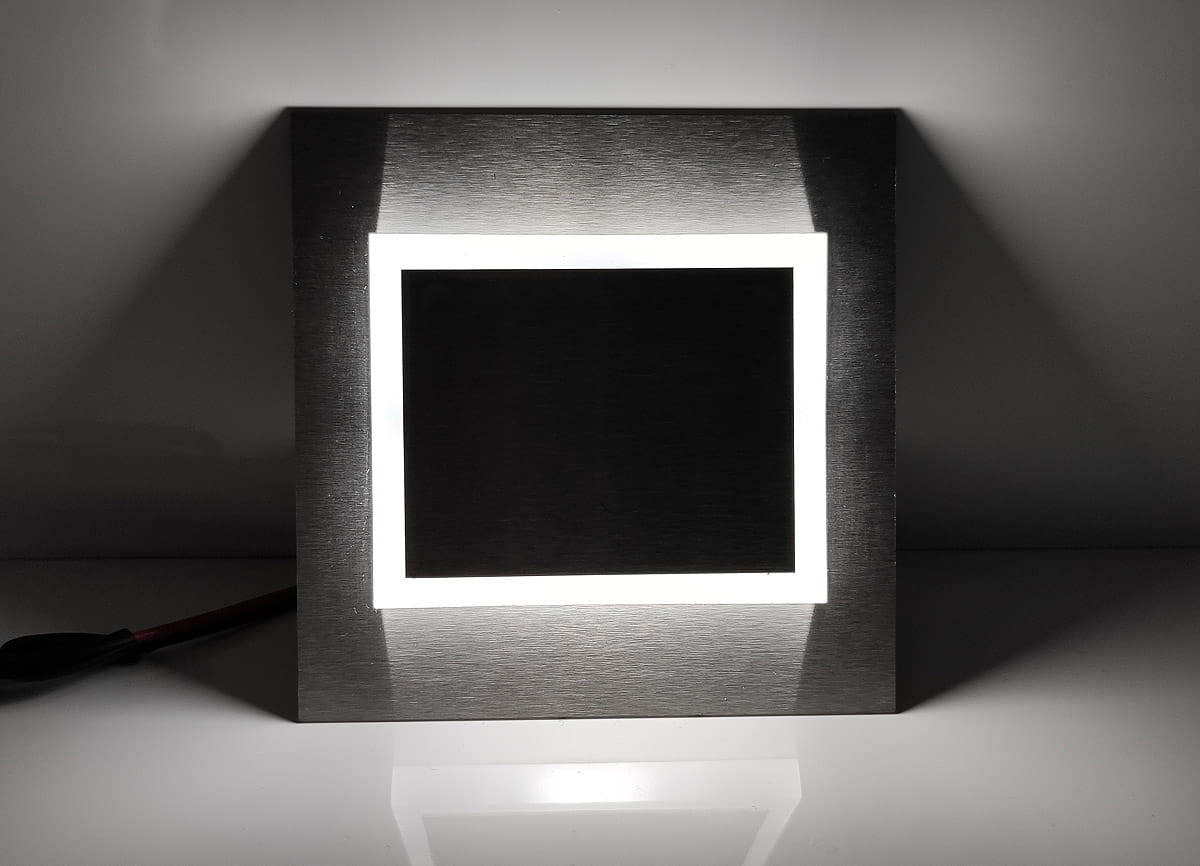 Oprawa schodowa Deco LED 230V w kolorze srebrnym, z zimnym światłem 6500K. Kwadratowy kształt z podświetlaną ramką, umieszczona frontalnie na jasnym tle. Widoczny kabel zasilający.