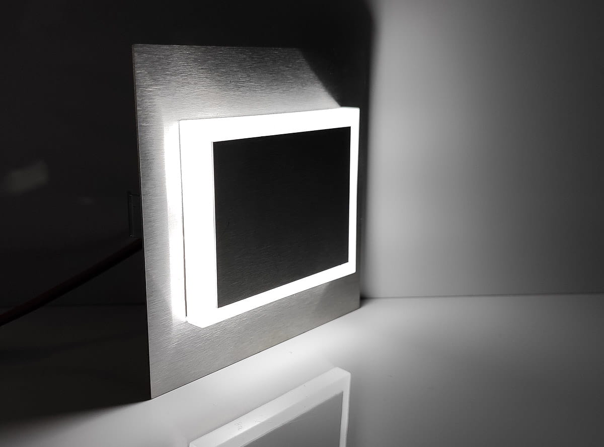 Oprawa schodowa Deco LED 230V w kształcie prostokąta, z metaliczną obudową i białym podświetleniem. Barwa zimna 6500K. Ujęcie z boku, z widocznym detalem świecącej krawędzi. Tło jednolite, podkreślające produkt.