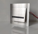 Oprawa schodowa Fero LED 230V w kolorze srebrnym, z podwyższonym, prostokątnym frontem i czarnym wcięciem, z podłączeniem elektrycznym z tyłu. Emituje ciepłe światło o temperaturze 3000K. Ujęcie z przodu, stojąca na białym tle.