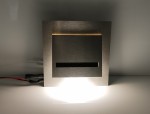 Oprawa schodowa Fero LED 230V w neutralnej barwie 4000K, wykonana z matowego metalu. Posiada prostokątny kształt z wycięciem oraz emitującą światło dolną część. Widoczne przewody zasilające w postaci czarno-czerwonych kabli.