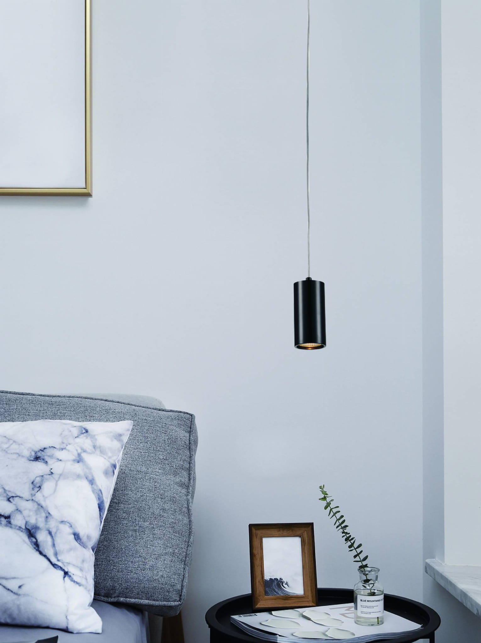 Lampka wisząca Kika S 85 w kolorze czarnym, o cylindrycznym kształcie, zawieszona na cienkim przewodzie. W tle szara kanapa z poduszką w marmurowy wzór oraz stolik z ramką i rośliną. Tło jasnoniebieskie.