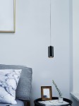 Lampka wisząca Kika S 85 w kolorze czarnym, o cylindrycznym kształcie, zawieszona na cienkim przewodzie. W tle szara kanapa z poduszką w marmurowy wzór oraz stolik z ramką i rośliną. Tło jasnoniebieskie.