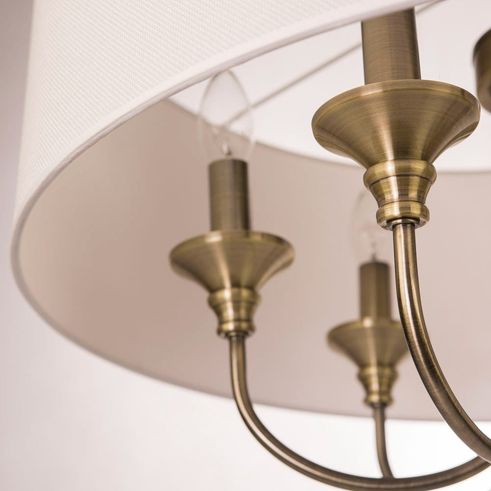Lampa wisząca Abu Dhabi 6-punktowa w kolorze patynowym. Klosz wykonany z białego materiału, z widocznymi metalowymi elementami w odcieniu patyny. Ujęcie zbliżeniowe ukazujące detale mocowania oraz kształt ramion.