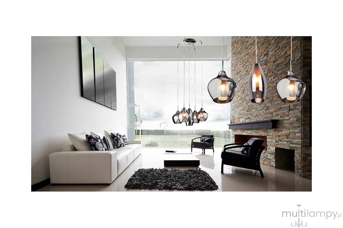 Lampa wisząca Amber Milano 1-punktowa w chromowanej obudowie, z przezroczystym kloszem. Produkt zawieszony w nowoczesnym wnętrzu, nad białą sofą z poduszkami, obok czarnych foteli i kominka. Widoczne ciepłe światło wewnątrz klosza.