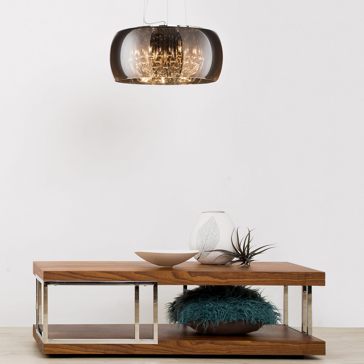 Lampa wisząca Crystal, 5-punktowa, w kolorze ciemnoszarym. Owalny klosz z przezroczystego tworzywa z centralnym źródłem światła, zawieszona na cienkich linkach. Ustawiona nad drewnianym stolikiem z metalowymi detalami.
