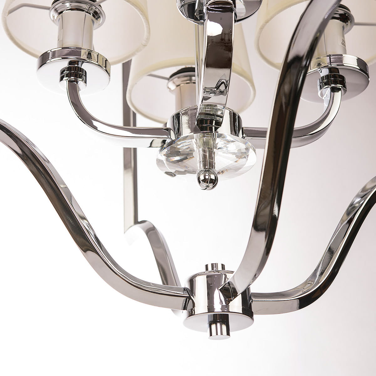 Lampa wisząca New York 4-punktowa, z metalowym, chromowanym stelażem. Widoczne cztery ramiona z kloszami w jasnym kolorze, centralny element z kryształem. Kąt ujęcia z dołu, ukazujący szczegóły konstrukcji. Tło jednolite, jasne.