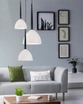 Lampa wisząca Eliza 1-punktowa w czarnym kolorze z białym kloszem, zawieszona na cienkim kablu. Umieszczona w nowoczesnym wnętrzu z szaro-skalnym tłem, obok kanapy i dekoracyjnych roślin.
