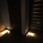 Lesel LED oprawa schodowa 002, umieszczona na ścianie, emituje miękkie światło w kolorze ciepłej bieli. Widoczna w aranżacji korytarza, oświetla podłogę. Oprawa ma minimalistyczny design, idealna do montażu w pobliżu drzwi.