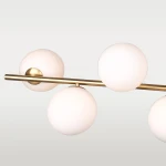 Lampa wisząca Cape Town 7-punktowa w kolorze mosiężnym z białymi, okrągłymi kloszami. Produkt zawieszony poziomo, z pięcioma kulistymi źródłami światła na metalowej rurze, na jasnym tle.