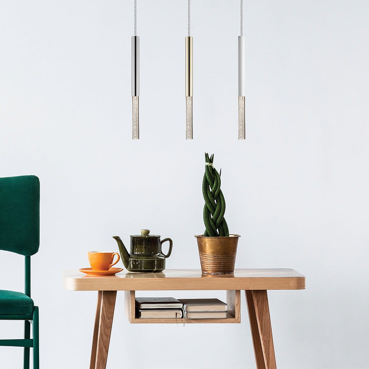 Wisząca lampa LED One, chromowana, z trzema prostokątnymi elementami z ozdobnym detalem. Zawieszona nad drewnianym stołem z zieloną rośliną i zastawą kawową. Tło jasne, minimalistyczne.