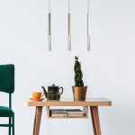 Wisząca lampa LED One, chromowana, z trzema prostokątnymi elementami z ozdobnym detalem. Zawieszona nad drewnianym stołem z zieloną rośliną i zastawą kawową. Tło jasne, minimalistyczne.