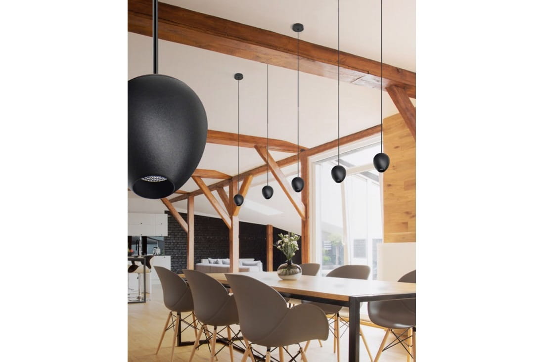 Czarna lampa wisząca Ovum LED z okrągłym kloszem, zawieszona na cienkim przewodzie. Widoczna w nowoczesnym wnętrzu z drewnianymi elementami. Lampa umieszczona nad stołem, w otoczeniu eleganckich krzeseł.