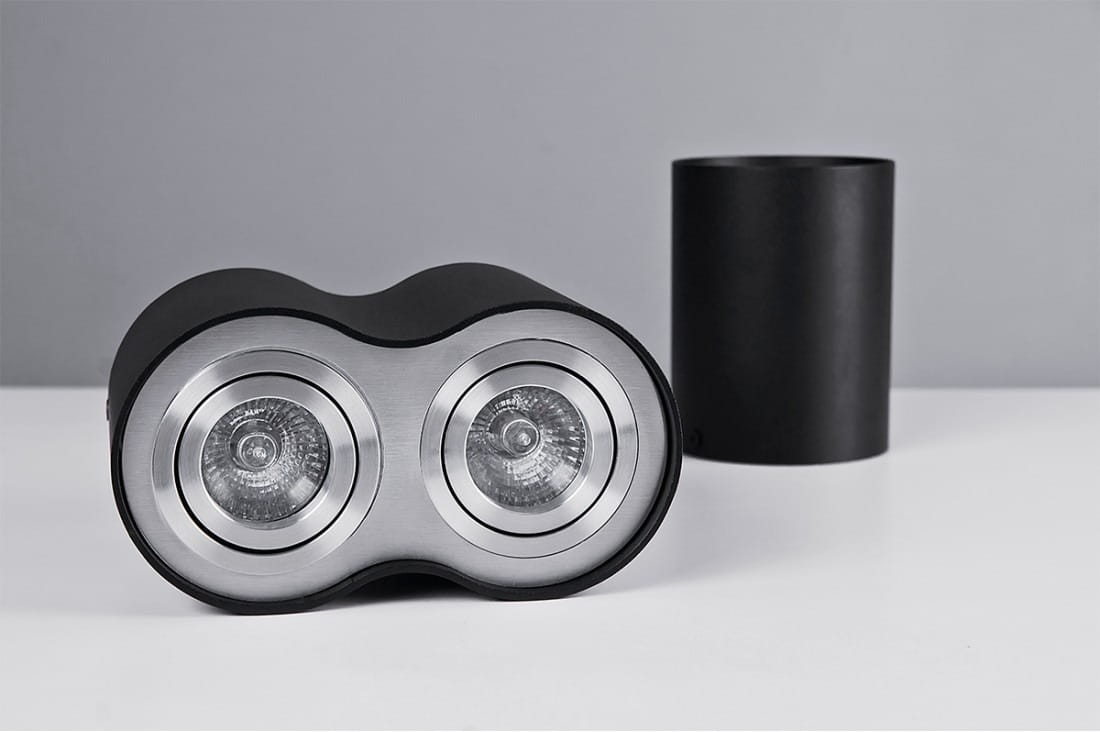 Czarna lampa sufitowa Bross z dwoma punktami świetlnymi, wykonana z aluminium. Ujęcie frontalne, lampy mają okrągłe, metalowe klosze z przezroczystymi żarówkami. W tle widoczny cylindryczny element w kolorze czarnym. Produkt leży na białym tle.