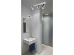 Lampa sufitowa Noemie IP44 w chromie, jedno punktowe oświetlenie. Zainstalowana na suficie w łazience, obok umywalki z lustrem. W tle widoczna szklana ścianka prysznicowa. Lampa z trzema ruchomymi reflektorami, które skierowane są w różne strony.