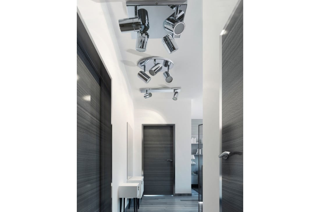 Noemie IP44 lampa sufitowa (spot) 2-punktowa w kolorze chrom. Ujęcie z przodu, zamontowana na suficie, z dwoma regulowanymi reflektorami. Tło stanowi jasny korytarz z drewnianymi drzwiami i minimalistycznym meblem.