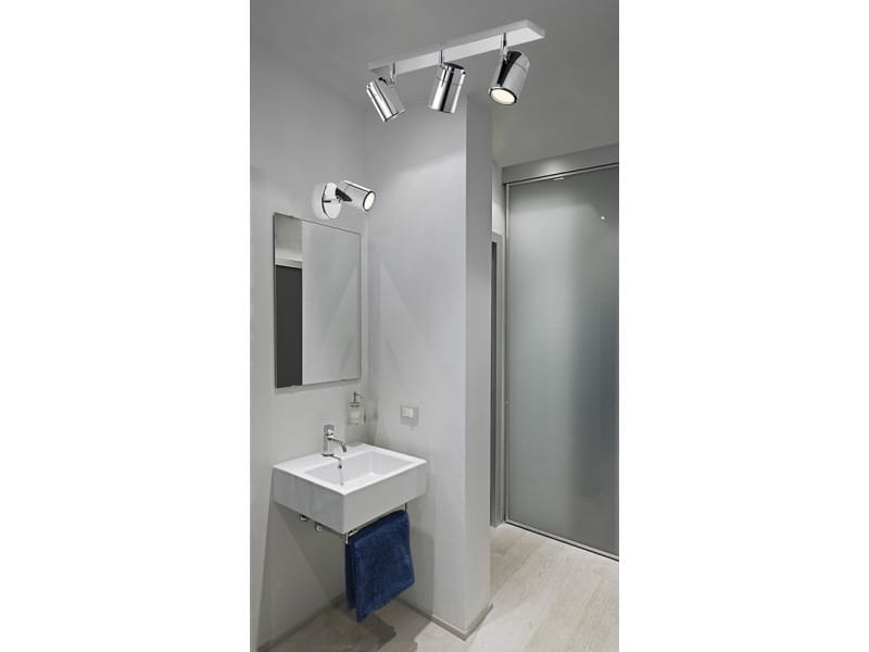 3-punktowa lampa sufitowa Noemie IP44 w chromowanym wykończeniu, zamontowana na suficie w łazience. Lampy mają cylindryczny kształt, emitując światło na lustro i umywalkę. W tle widoczna szklana ścianka prysznicowa i ręcznik w niebieskim kolorze.