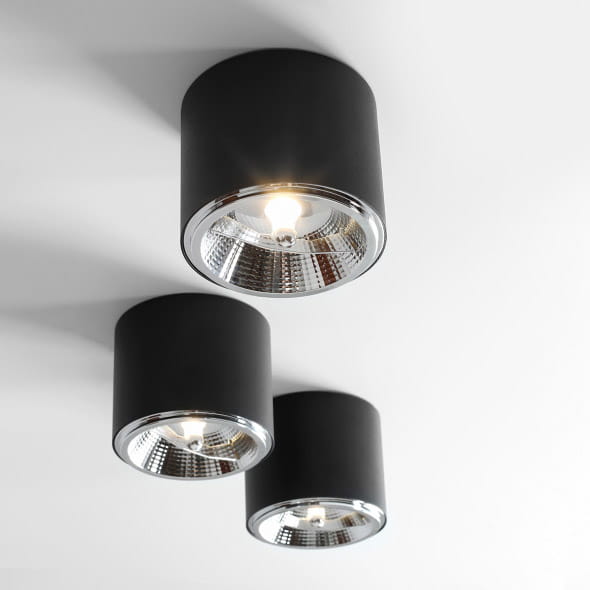 Czarna lampa sufitowa spot 1-punktowa, cylindryczny kształt z metalicznym wykończeniem, zamontowana na białym suficie. Wyraźne źródło światła widoczne w środku reflektora, trzy lampy w linii. Minimalistyczny design, idealna do nowoczesnych wnętrz.