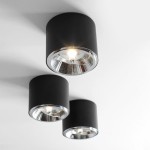 Czarna lampa sufitowa spot 1-punktowa, cylindryczny kształt z metalicznym wykończeniem, zamontowana na białym suficie. Wyraźne źródło światła widoczne w środku reflektora, trzy lampy w linii. Minimalistyczny design, idealna do nowoczesnych wnętrz.
