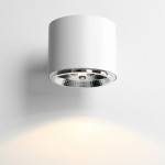 Biały kinkiet 1-punktowy Bot 1046C o cylindrycznym kształcie, z metalicznym wykończeniem na spodzie klosza. Zamontowany na ścianie, emituje ciepłe światło w dół. Gładka faktura, minimalistyczny design, idealny do nowoczesnych wnętrz.