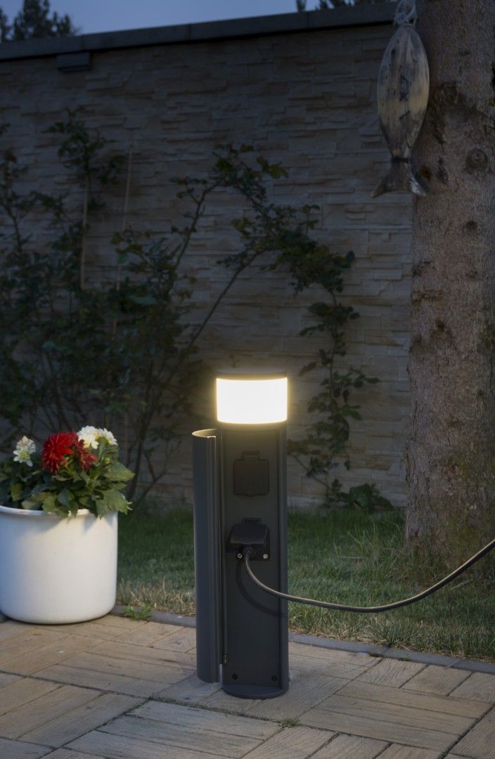 Lampa stojąca ogrodowa Elborn w kolorze szarym, z okrągłym kloszem emitującym ciepłe światło. Posiada wbudowane gniazda elektryczne, umieszczona na płaskiej powierzchni, w tle widoczne kwiaty w donicy oraz mur.
