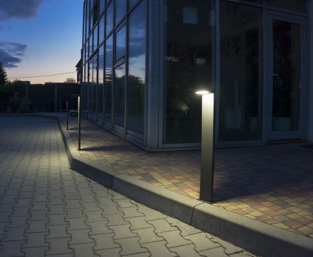 Lampa stojąca ogrodowa Karo LED H78 w kolorze szarym, o prostokątnym kształcie, umieszczona na krawędzi chodnika. Źródło światła znajduje się na górze, oświetlając przestrzeń wokół. Tło to nowoczesny budynek ze szklanymi ścianami.