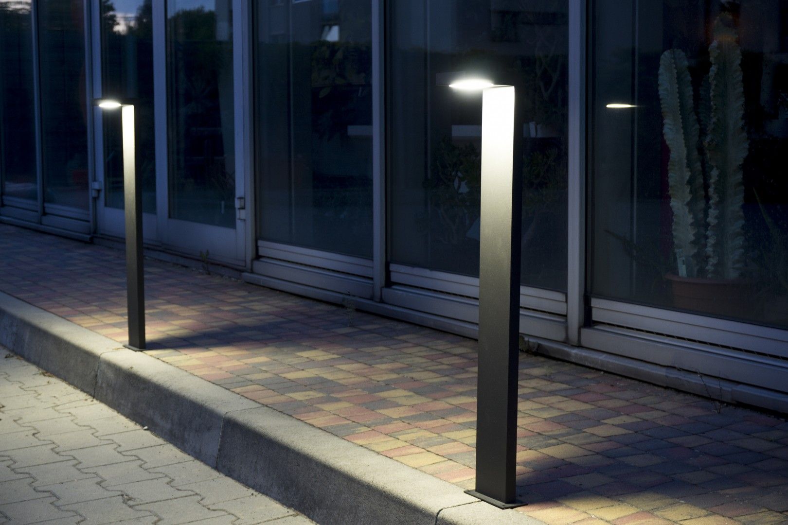 Lampa stojąca ogrodowa Karo LED H78, 1-punktowa, o prostokątnym kształcie, wykonana z metalu w kolorze czarnym. Ujęcie frontalne, lampa emituje światło w górę. Znajduje się na chodniku, w tle przeszklona ściana budynku oraz roślinność.