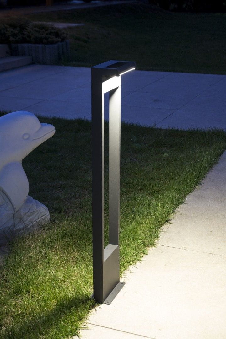 Lampa stojąca ogrodowa Rota LED H78 w kolorze ciemnoszarym, o nowoczesnym, prostokątnym kształcie. Widok z boku, z włączonym oświetleniem, na tle trawnika i chodnika, obok rzeźby ptaka. Dostosowana do oświetlenia przestrzeni zewnętrznej.