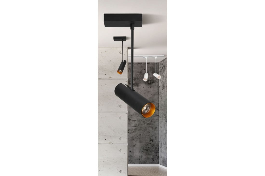 Czarna lampa sufitowa (spot) Lens Square z metalowym cylindrem i złotym wnętrzem, zawieszona pod kątem. Montaż na suficie, widoczny także drugi, mniejszy spot w tle. Tło w odcieniach szarości z betonowymi ścianami.