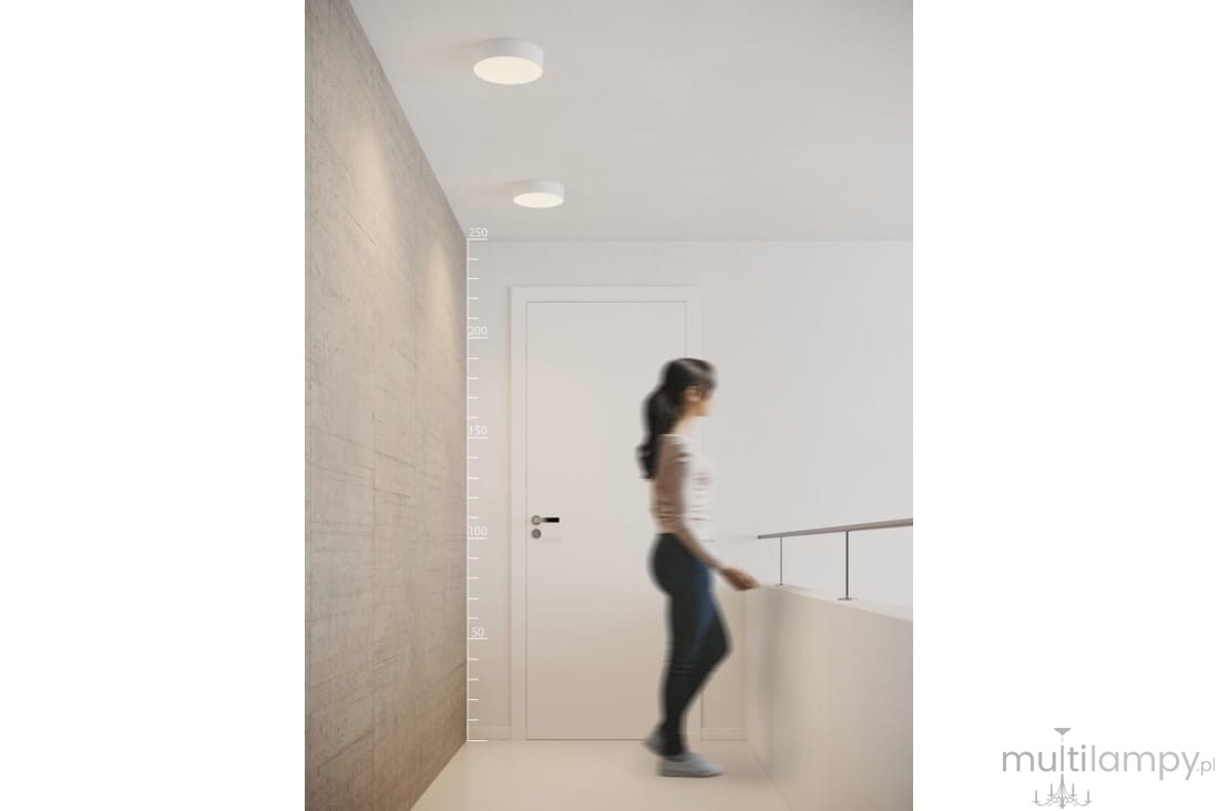 Plafon LED Monza R 22 4000K w kolorze białym, umieszczony na suficie w korytarzu. Oświetlenie jednolitym, okrągłym kształcie. W tle widoczna biała ściana i drzwi, a także postać kobiety przechodzącej obok balustrady.