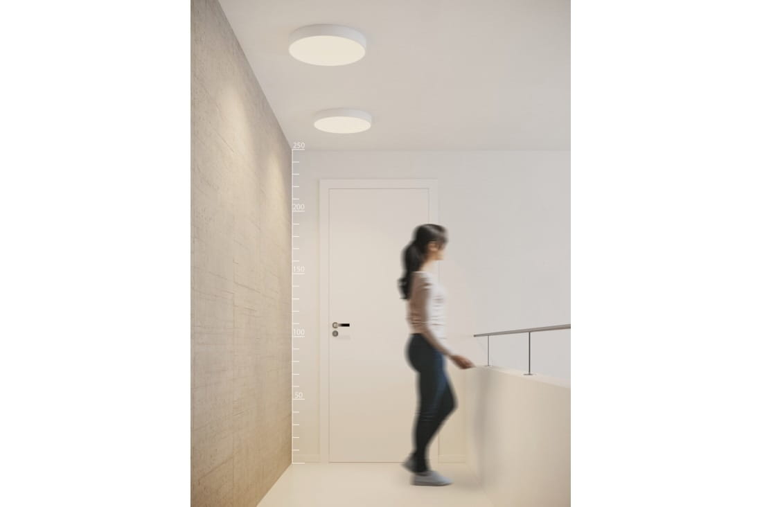 Plafon Monza R 40 3000K w kolorze białym, zamontowany na suficie. Okrągły kształt, minimalistyczny design. Widoczna osoba przechodzi w tle, w nowoczesnym wnętrzu z jasnymi ścianami. Tło proste, podkreślające produkt.