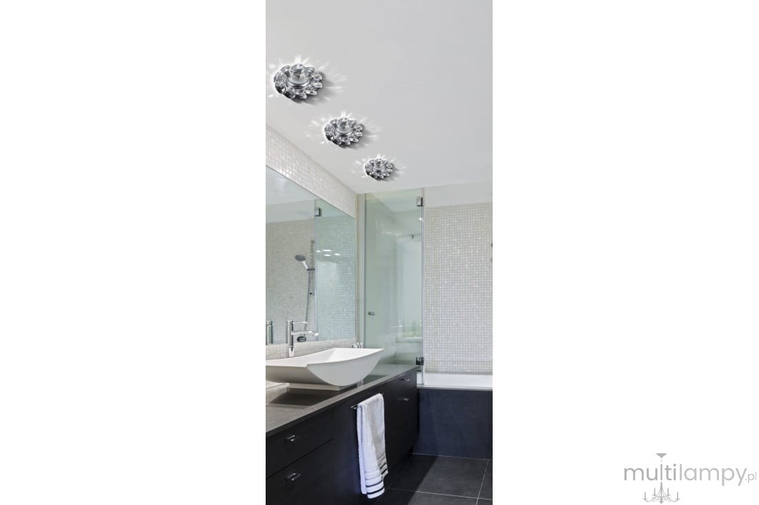Oprawa sufitowa Erika 1-punktowa w kolorze chromu, zamontowana w łazience. Widoczna z góry, elegancka forma z dekoracyjnym wzorem. Tło jasne, minimalistyczne wnętrze z lustrem, umywalką i ciemnymi szafkami.
