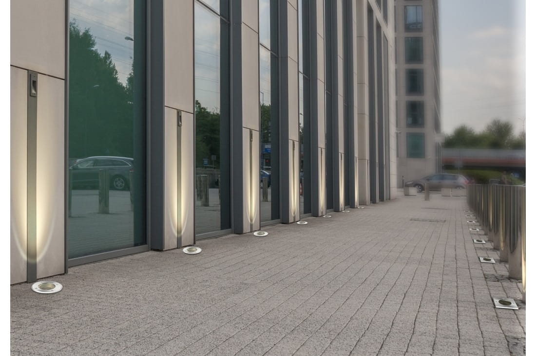 Oprawa dogruntowa Hyde Square 1-punktowa w kolorze chrom, umieszczona na nawierzchni przed budynkiem. Widoczna w pozycji leżącej, z okrągłą, metaliczną obudową i delikatnym oświetleniem wokół. Tło stanowi nowoczesna fasada budynku z dużymi oknami.
