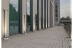 Oprawa dogruntowa Hyde Square 1-punktowa w kolorze chrom, umieszczona na nawierzchni przed budynkiem. Widoczna w pozycji leżącej, z okrągłą, metaliczną obudową i delikatnym oświetleniem wokół. Tło stanowi nowoczesna fasada budynku z dużymi oknami.
