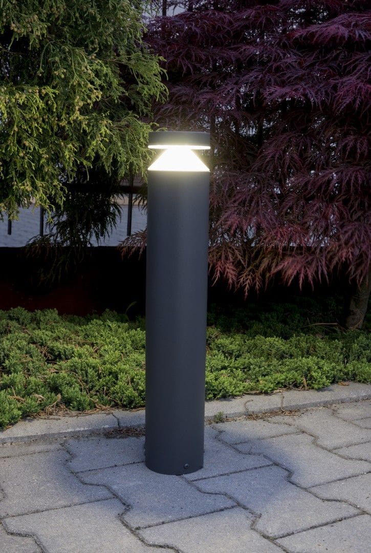 Lampa stojąca ogrodowa Delta LED H50, w kształcie cylindrycznym, w kolorze czarnym. Posiada górną, szklaną osłonę emitującą światło. Ujęcie frontalne, lampa ustawiona na betonowej nawierzchni, w otoczeniu zieleni.