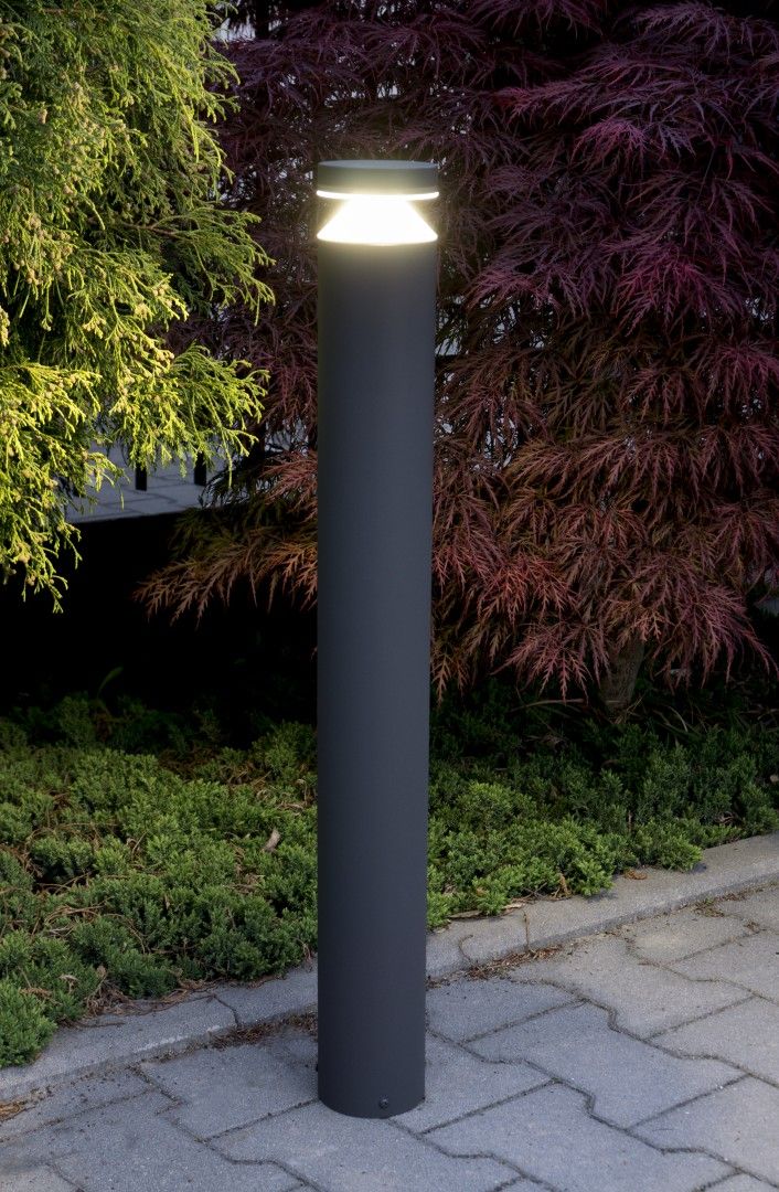 Lampa stojąca ogrodowa Delta LED H75 w kształcie cylindrycznym, wykonana z matowego, ciemnoszarego metalu. Posiada źródło światła na górze, emitujące jasne światło, umieszczona na kostce brukowej w otoczeniu zieleni.