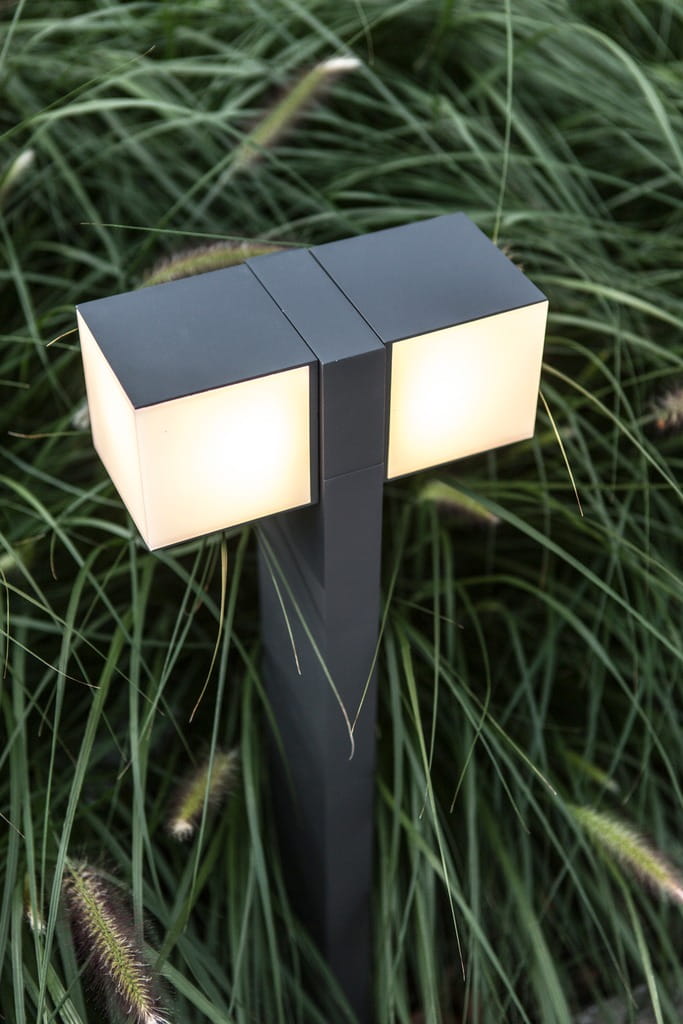 Lampa stojąca ogrodowa Cuba LED H75 w ciemnoszarej obudowie, z regulowanymi dwoma punktami świetlnymi. Ujęcie z boku, wśród zielonej trawy. Prostokątne klosze emitujące miękkie światło.