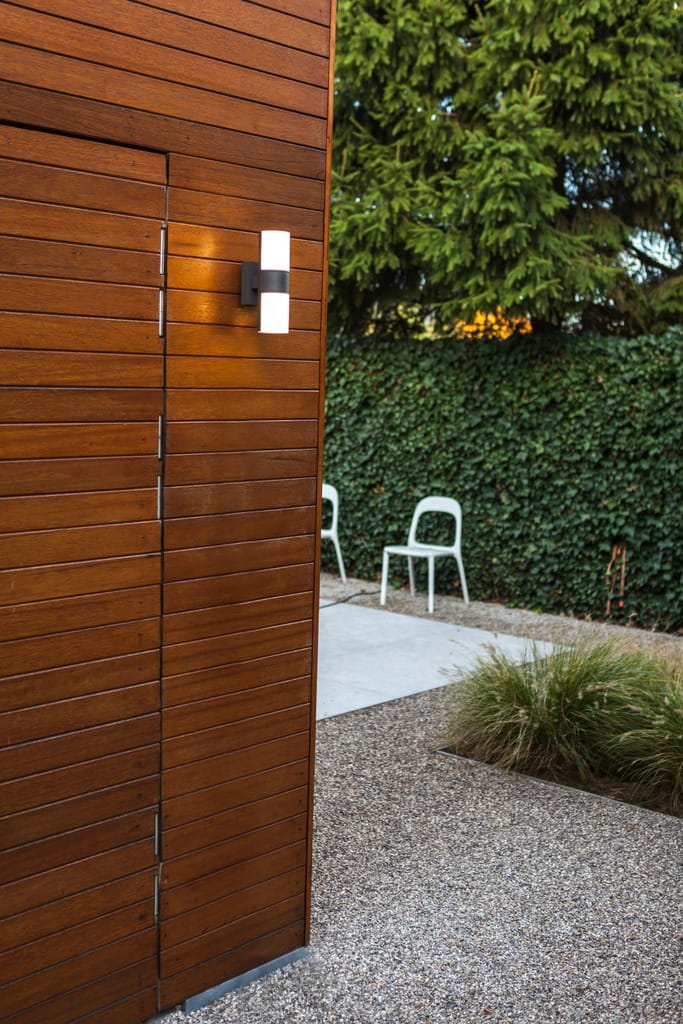 Regulowany kinkiet elewacyjny Cyra LED w ciemnoszarym kolorze, zamontowany na drewnianej ścianie. Oświetlenie 2-punktowe z białymi kloszami, otoczone zielenią i kamienistym podłożem, w tle widoczna biała krzesła.