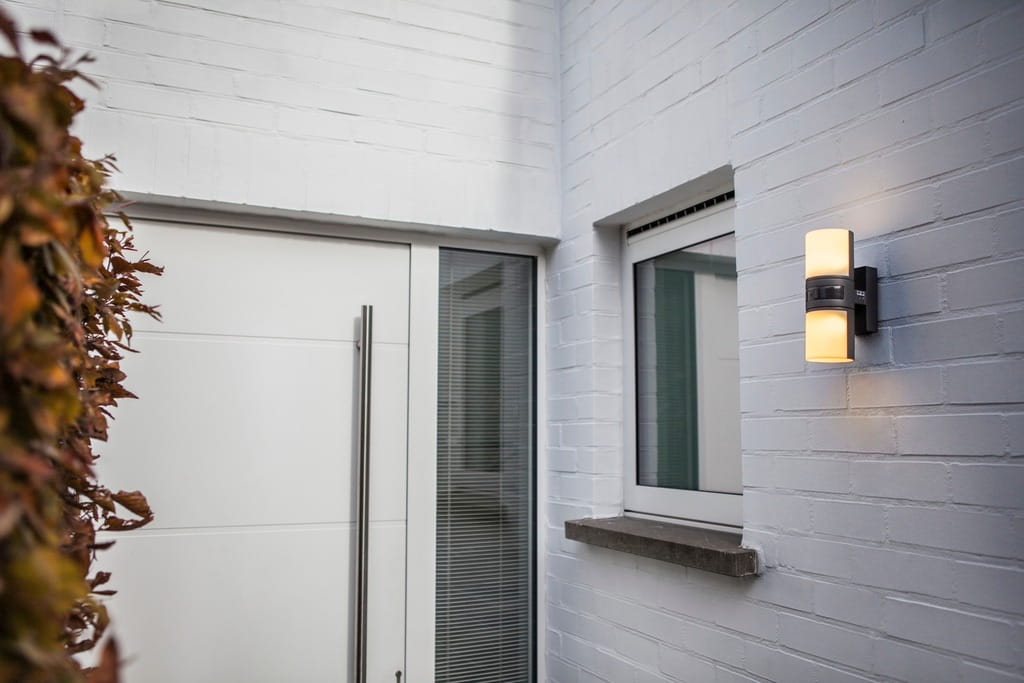 Cyra LED kinkiet elewacyjny 2-punktowy w ciemnoszarym kolorze, zamontowany na ścianie, emituje ciepłe światło. W tle widoczne białe drzwi i okno, z roślinnością w pobliżu. Kinkiet wyposażony w czujnik ruchu.