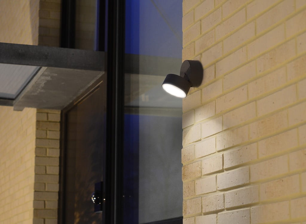 Kinkiet elewacyjny Trumpet LED 1-punktowy w ciemnoszarym kolorze, zamontowany na jasnej ceglanej ścianie. Lampa z regulowanym kątem świecenia, emitująca światło na powierzchnię pod nią. W tle widoczna szklana fasada budynku.
