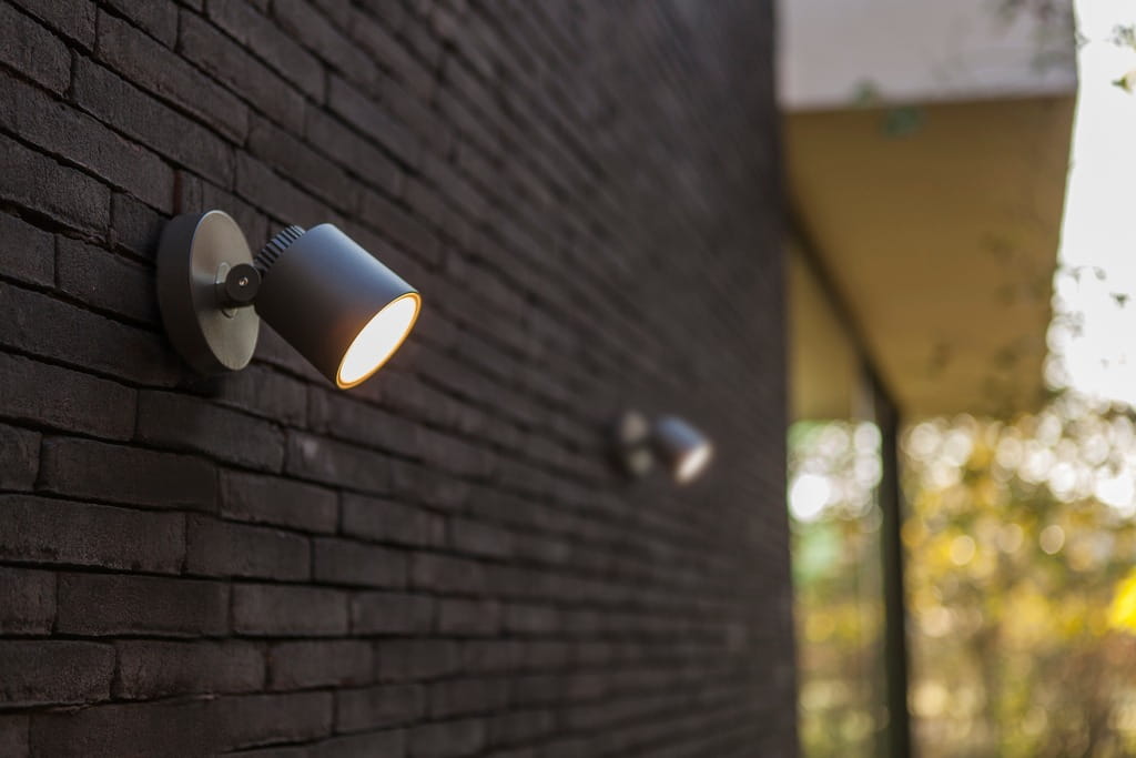 Ciemnoszary kinkiet elewacyjny Explorer LED, 1-punktowy, z regulowanym kloszem, zamontowany na ścianie z cegły. Klosz skierowany w dół, emitujący światło. Tło z zielonymi akcentami w oddali.