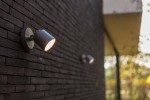 Ciemnoszary kinkiet elewacyjny Explorer LED, 1-punktowy, z regulowanym kloszem, zamontowany na ścianie z cegły. Klosz skierowany w dół, emitujący światło. Tło z zielonymi akcentami w oddali.