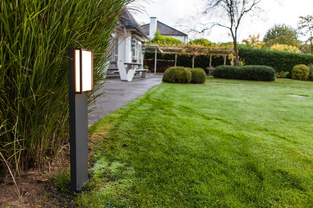 Lampa stojąca ogrodowa Qubo LED H75 w ciemnoszarym kolorze, prostokątna, z białym kloszem. Ustawiona na trawie, w otoczeniu zieleni, z widokiem na podjazd i roślinność.