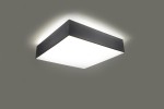 Lampa sufitowa Horus 25, 1-punktowa, w kolorze szarym. Prosta, kwadratowa forma z matowym białym kloszem, zamontowana na suficie, emitująca delikatne światło. Tło w odcieniu szaro-beżowym.