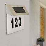 Podświetlany numer domu w formie kinkietu, z metalową obudową i panelami słonecznymi, montowany na ścianie. Wyraźny numer 123 na białym tle, umieszczony pod osłoną. Czujnik zmierzchu automatycznie włącza światło po zapadnięciu zmroku.