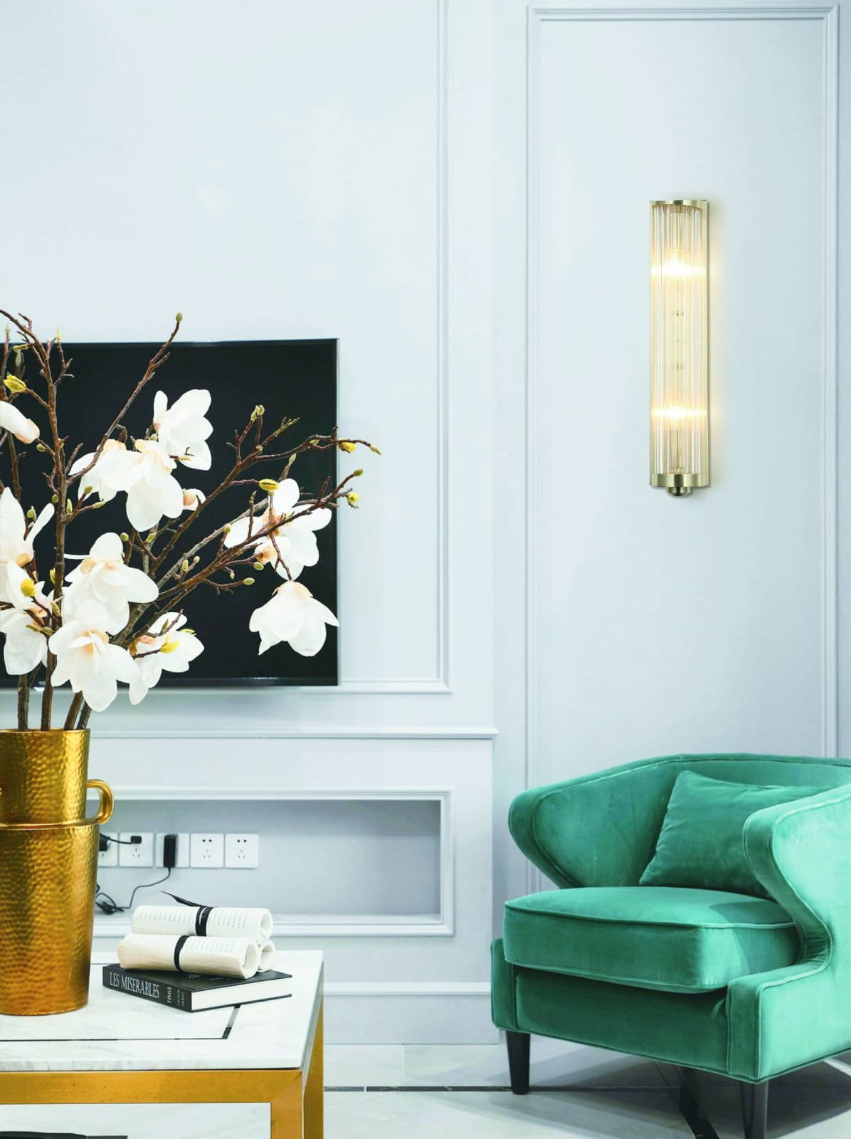 Kinkiet Fiatto Parette Cromo w kolorze chromu, o prostokątnym kształcie, z przezroczystymi, poziomymi prętami. Zawieszony na jasnej ścianie obok zielonego fotela, w aranżacji z białymi kwiatami w złotym wazonie.