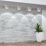 Biała lampa sufitowa kierunkowa Hadar 1xGU10, o prostokątnym kształcie, zamontowana na suficie. Oświetlenie skierowane na ścianę z cegły, w tle widoczna roślina w białym doniczce. Prosta, nowoczesna forma, idealna do aranżacji wnętrz.