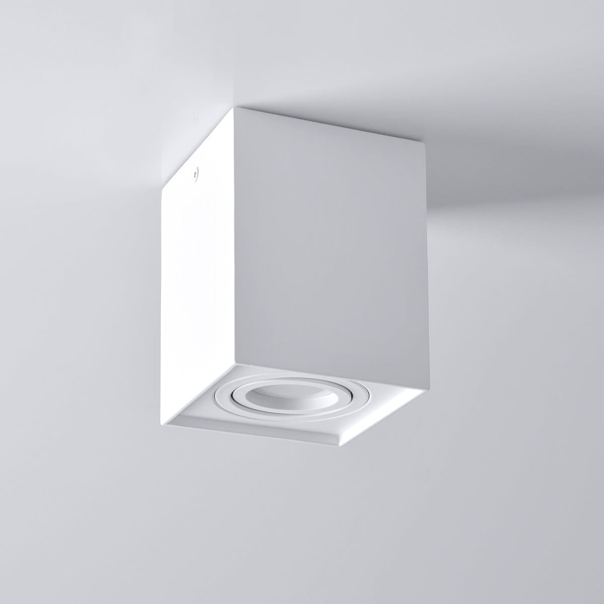 Lampa sufitowa Hadar w kolorze białym, z kwadratowym korpusem. Posiada jedno gniazdo GU10, zamontowana na suficie. Zbliżenie na okrągły element źródła światła, minimalistyczny design, idealna do nowoczesnych wnętrz.