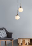 Lampa wisząca Erich 1-punktowa w kolorze mosiądzu z okrągłym, matowym kloszem. Produkt zawieszony na cienkim przewodzie, z widocznym uchwytem. W tle szara ściana, obok szary fotel z poduszką i stolik z drewnianymi dekorami.